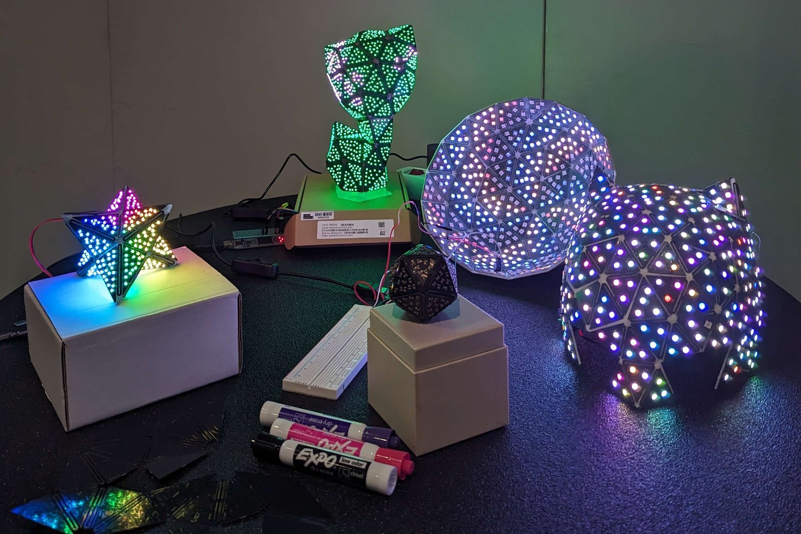 Five fabricated PCBend models lit with RGB LEDs, next to unfolded PCB panels